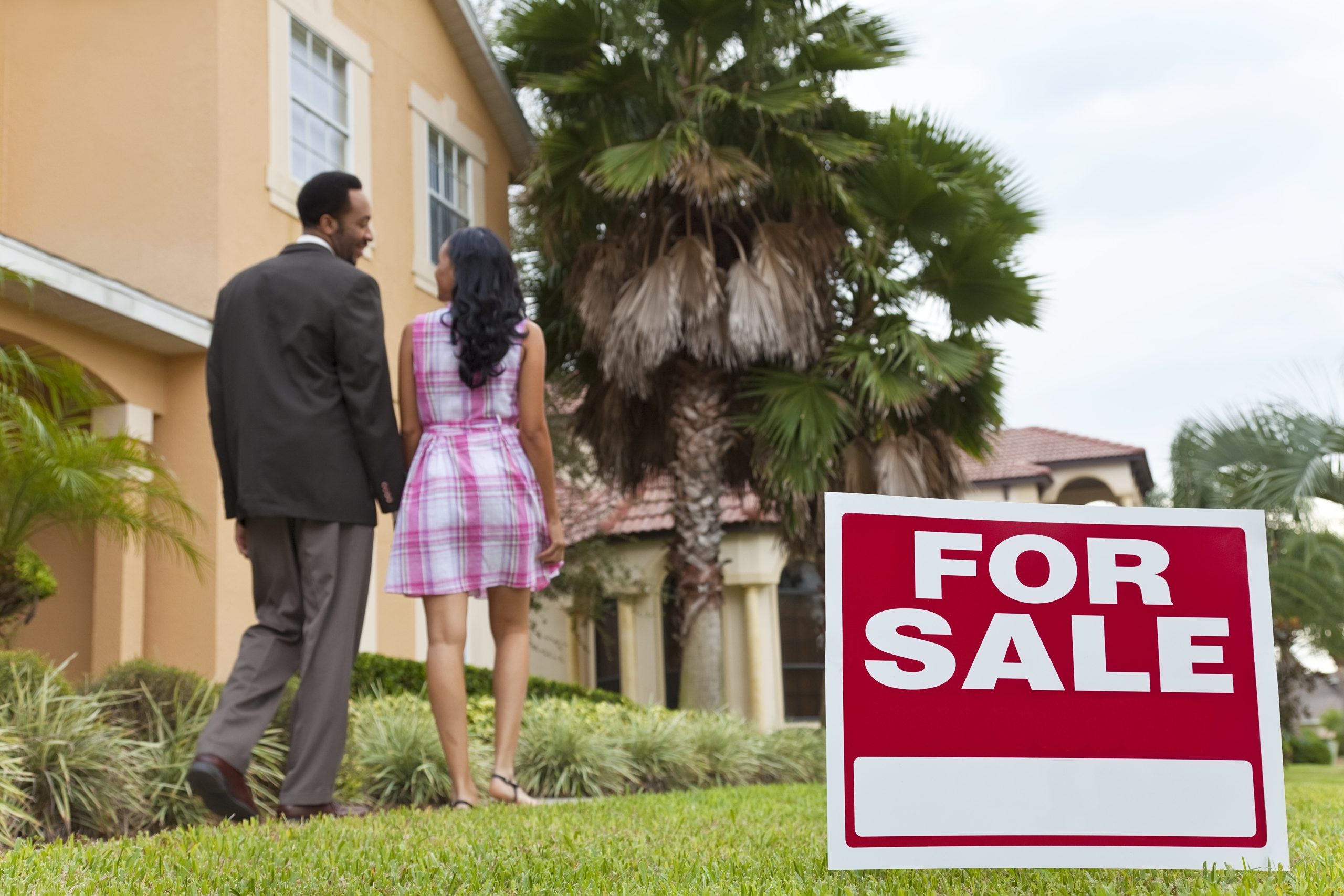 A happy African American man and woman couple house hunting outside a large house with a For Sale sign. The focus is on the sign.